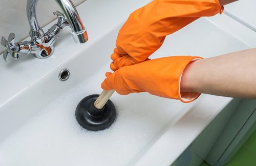 Một người đeo găng tay bảo hộ màu cam thông bồn rửa bị tắc bằng pít-tông hoặc bơm cao su.  Cận cảnh, tập trung chọn lọc.