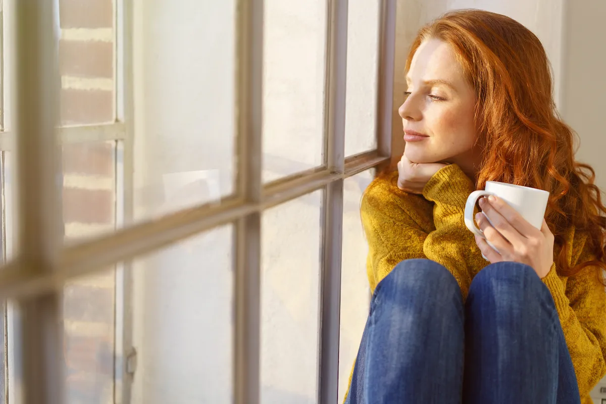Pretty redhead woman savoring her mug of hot coffee as she sits on a window sill with eyes closed in contentment