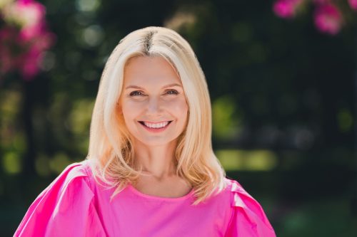 Portrait of a smiling middle-aged blonde woman wearing a bright pink shirt while standing outside