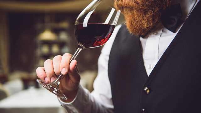 A close up of a person with a beard sniffing from a glass of red wine