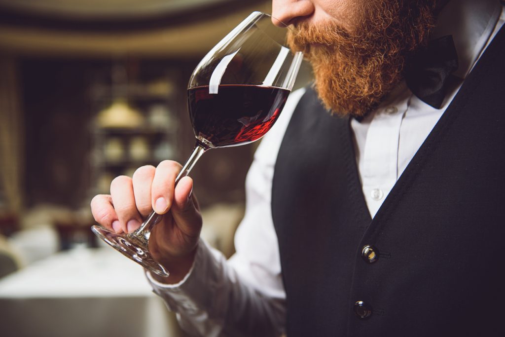 A close up of a person with a beard sniffing from a glass of red wine