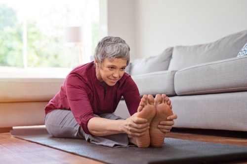 Beautiful senior woman doing stretching exercise while sitting on yoga mat at home. Mature woman exercising in sportswear by stretching forward to touch toes