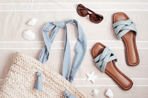 Summer vacations flat lay with beach accessories. Blue leather sandals, sunglasses, beach bag, and seashells over beach towel background