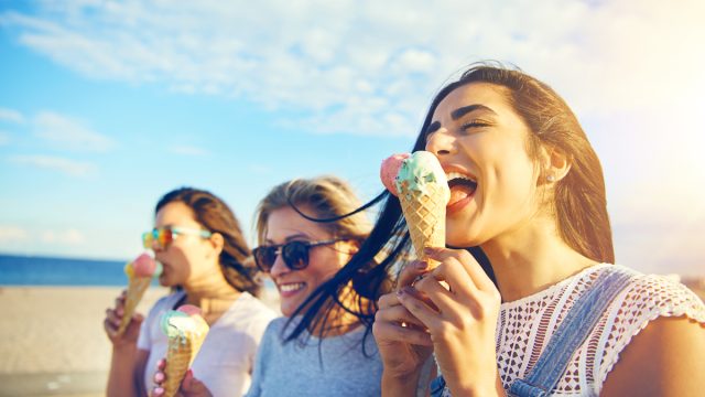 Three young woman eating ice cream cones at the seaside as they stroll along a waterfront promenade