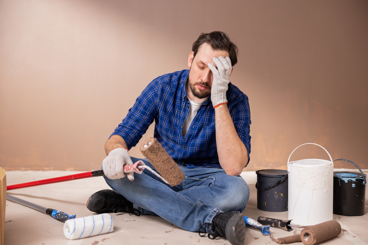 An upset-looking man sitting on the floor with his head in his hands while surrounded by painting supplies