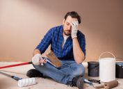 An upset-looking man sitting on the floor with his head in his hands while surrounded by painting supplies