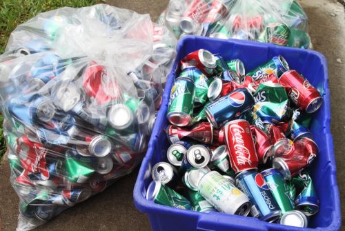 Aluminum soda cans are being stored for recycling. Two bags and a bin of cans contain a variety of brand sodas, including Coca Cola, Pepsi, Sprite, Dr Pepper, Mountain Dew and Mist.