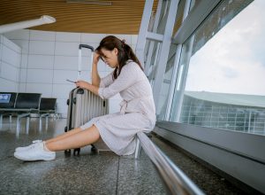 A woman sitting on the ground next to her suitcase in the airport looking disappointed or upset