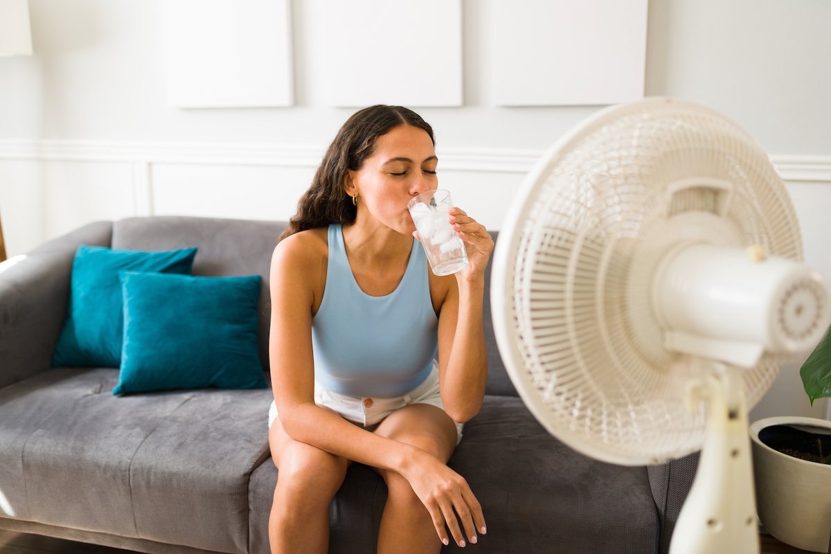 Woman sitting on her couch drinking a glass of water with a fan pointed at her