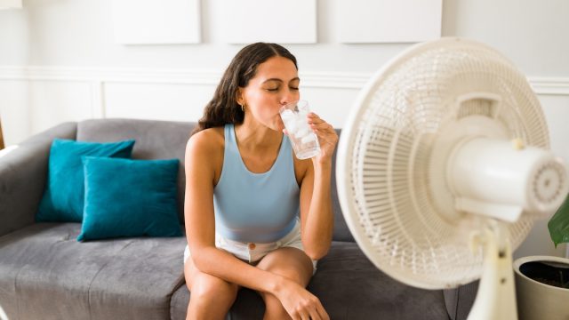 Woman sitting on her couch drinking a glass of water with a fan pointed at her