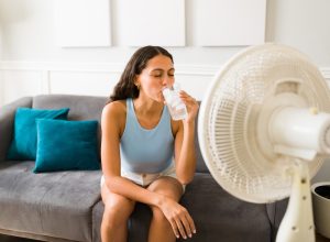 Woman sitting on her couch drinking a glass of water with a fan pointed at her