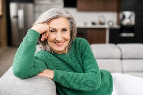 A smiling older woman with gray hair wearing a green sweater sits on the couch with her head leaning on her hand