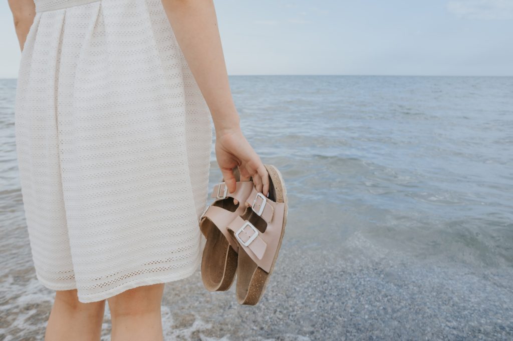 Người phụ nữ mặc đồ trắng bạc màu đứng gần biển cầm đôi dép Birkenstock màu be