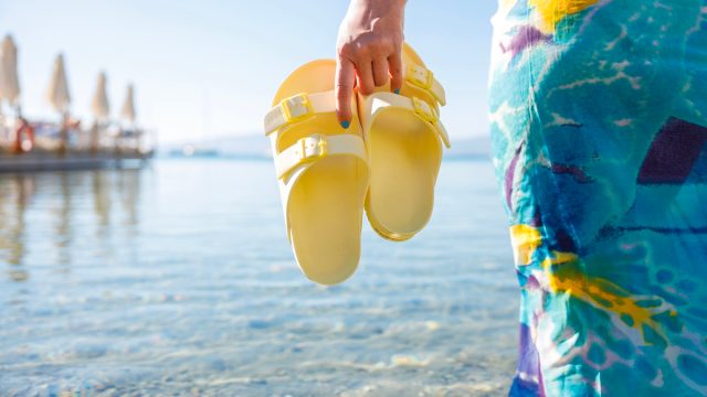 Cropped image of a woman in a blue dress standing in the ocean holding a pair of yellow Birkenstock-style sandals