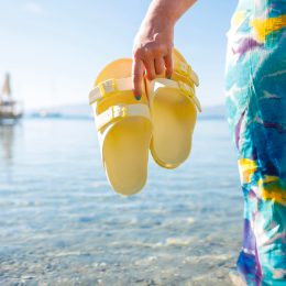 Cropped image of a woman in a blue dress standing in the ocean holding a pair of yellow Birkenstock-style sandals