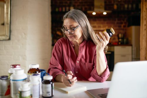 Portrait of middle aged woman in eyeglasses making research, sitting in front of open laptop, handwriting in copybook, studying dietary supplements, holding bottle of vitamins