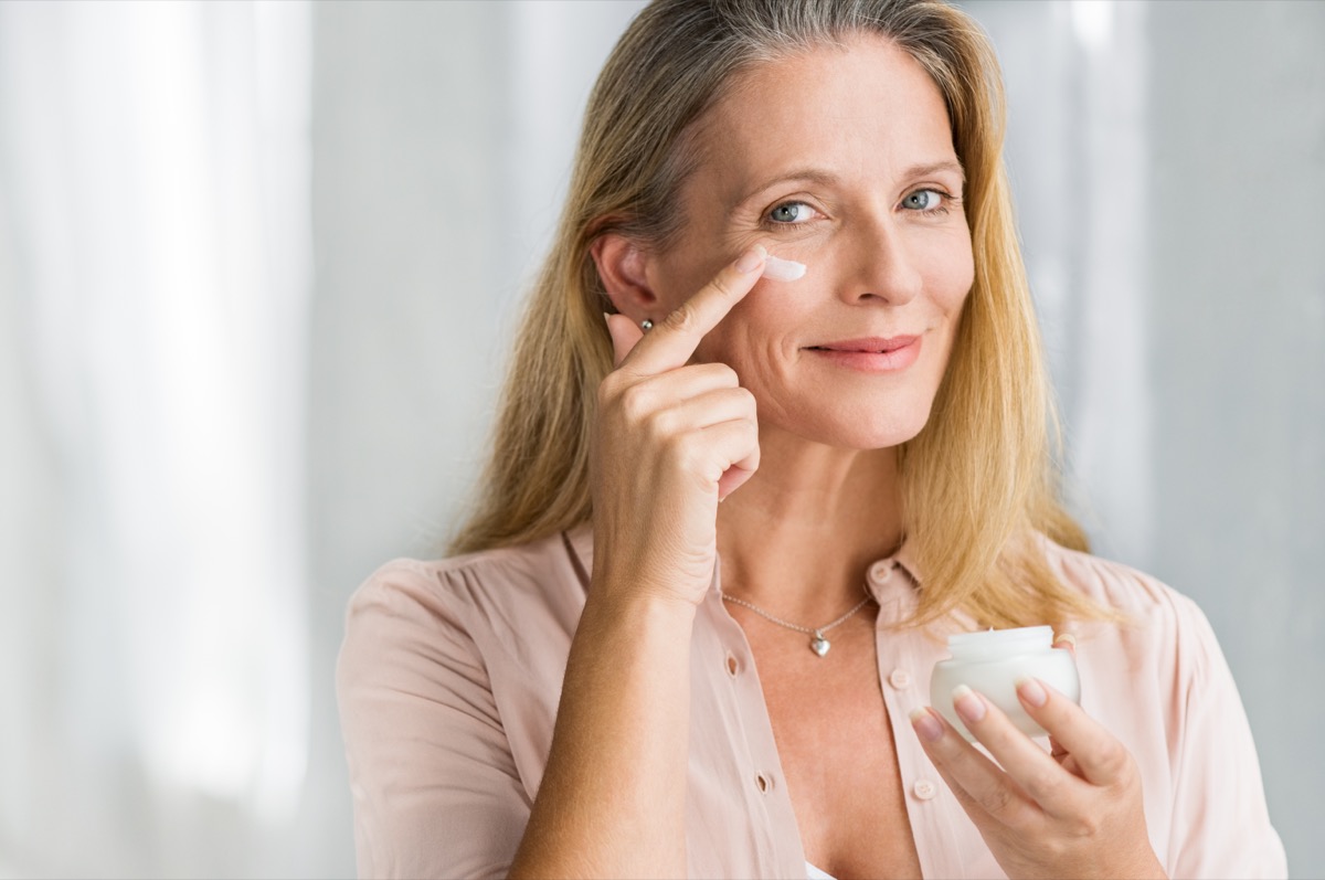 Smiling senior woman applying anti-aging lotion to remove dark circles under eyes. Happy mature woman using cosmetic cream to hide wrinkles below eyes. Lady using day moisturizer.