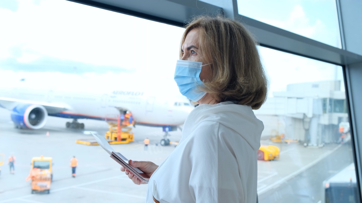 adult Female in a protective mask stands at the window in the airport terminal awaiting the departure of a flight due to travel restrictions due to the coronavirus pandemic, a senior aged 50-55 holds