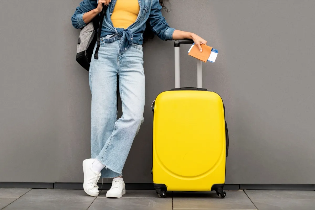 Unrecognizable woman in stylish casual outfit traveller standing over grey background, carrying yellow luggage and backpack, holding passport and flight tickets, cropped, copy space