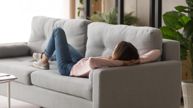 Millennial girl lying and relaxing on comfortable couch after hard work day. Young calm female student having rest, daydreaming, refreshing in living room at home, lazy weekend peace of mind concept.