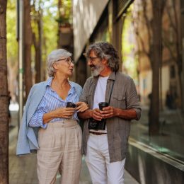 A cheerful senior couple is outside in the city walking on the street, socializing and drinking coffee.