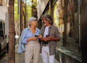 A cheerful senior couple is outside in the city walking on the street, socializing and drinking coffee.