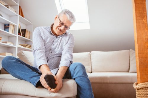Senior man suffering with foot cramp on sofa in living room at home.