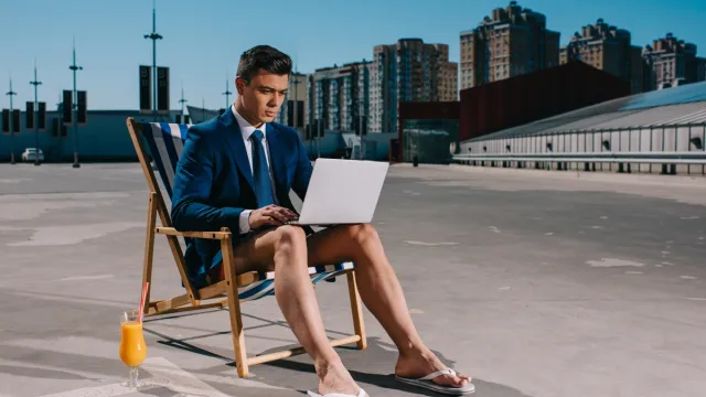 young businessman in shorts and jacket working with laptop while sitting on sun lounger