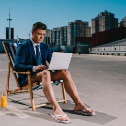 young businessman in shorts and jacket working with laptop while sitting on sun lounger