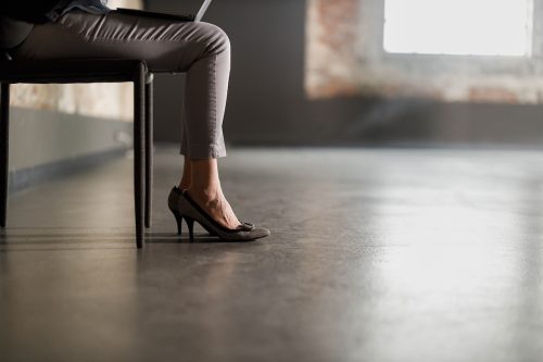 Unrecognizable female entrepreneur wearing heels and sitting on a chair with laptop on her lap in an empty office space.