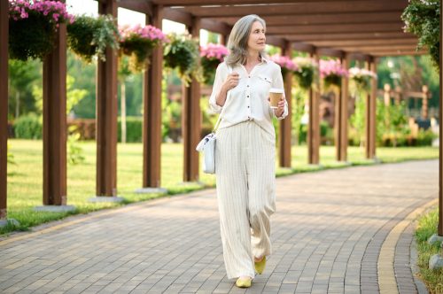 A mature woman wearing a beige linen outfit walks in a garden with a coffee