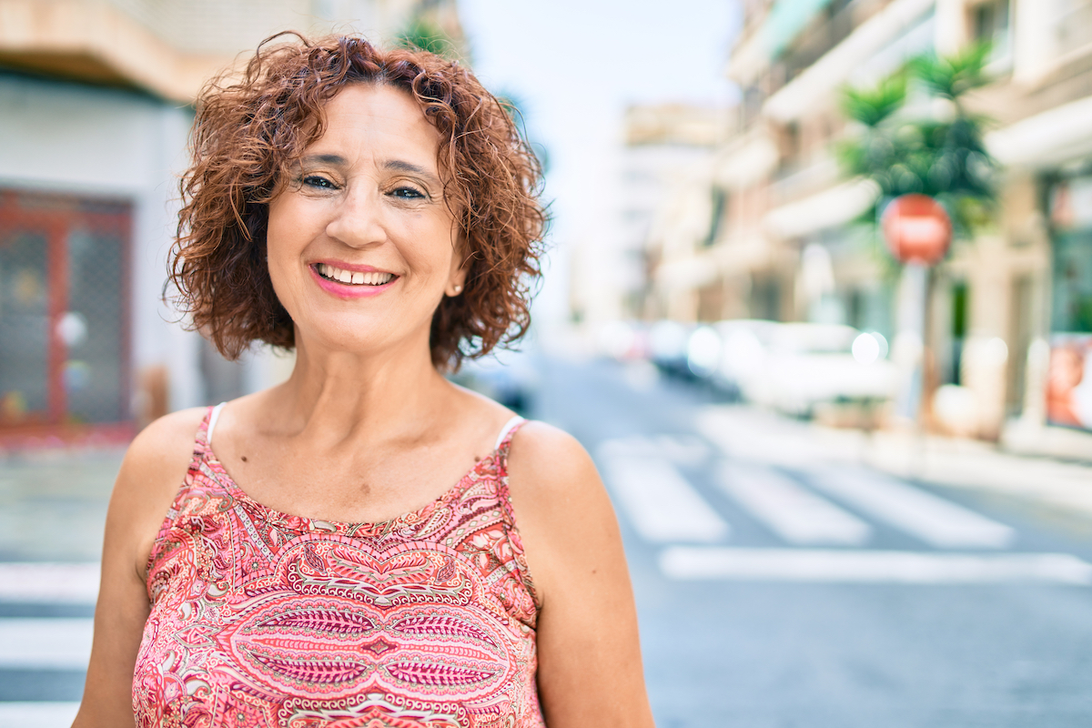 A mature woman with red curly hair smiling on a city street, wearing a red paisley tank top.
