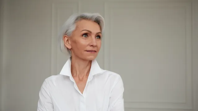 Portrait of an adult elderly self-confident woman with white hair looking awey into distance in white shirt hands crossed background wall