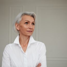 Portrait of an adult elderly self-confident woman with white hair looking awey into distance in white shirt hands crossed background wall