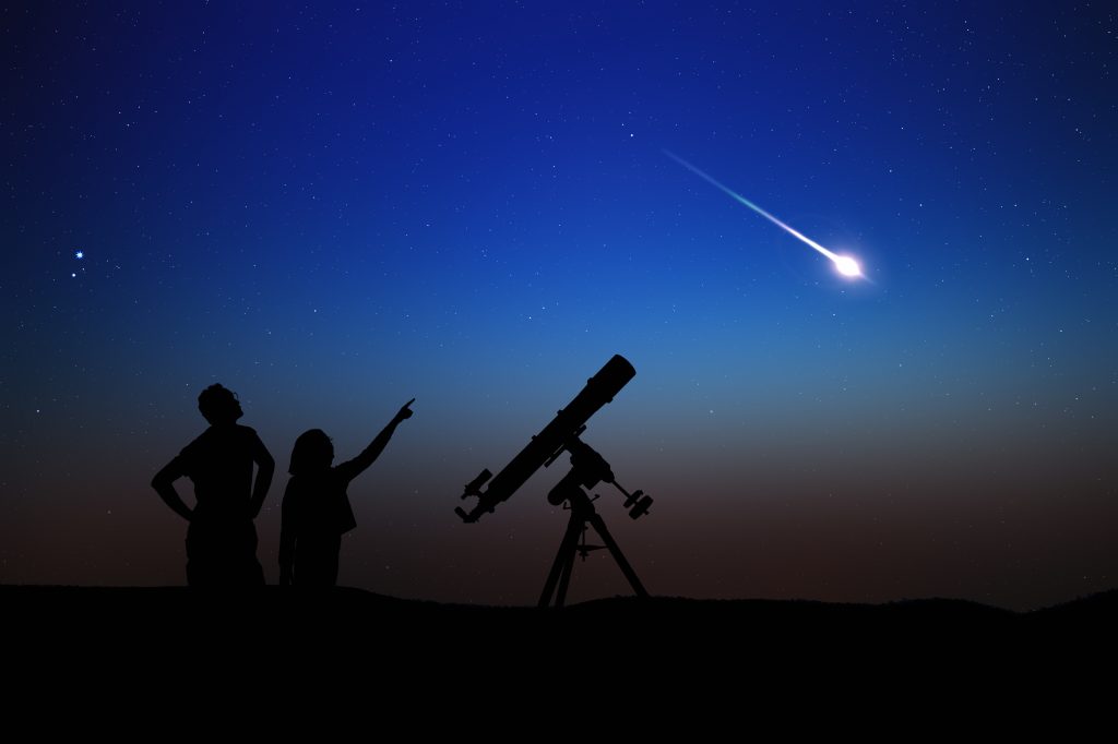 A silhouette of a parent and child watching a shooting star next to their telescope