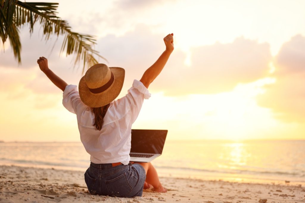 woman with arms raised sitting with laptop on her legs on a beach at sunset
