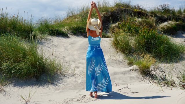 Rear view of a woman standing among sand dunes, wearing a long blue dress and a sun hat