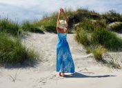 Rear view of a woman standing among sand dunes, wearing a long blue dress and a sun hat
