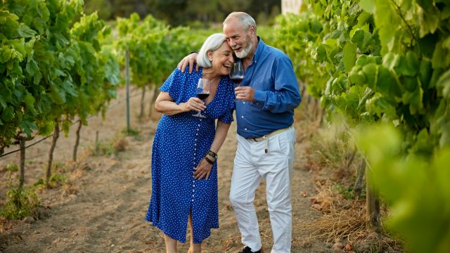 Senior couple, both wearing blue, hugging and laughing while drinking red wine in a vineyard.