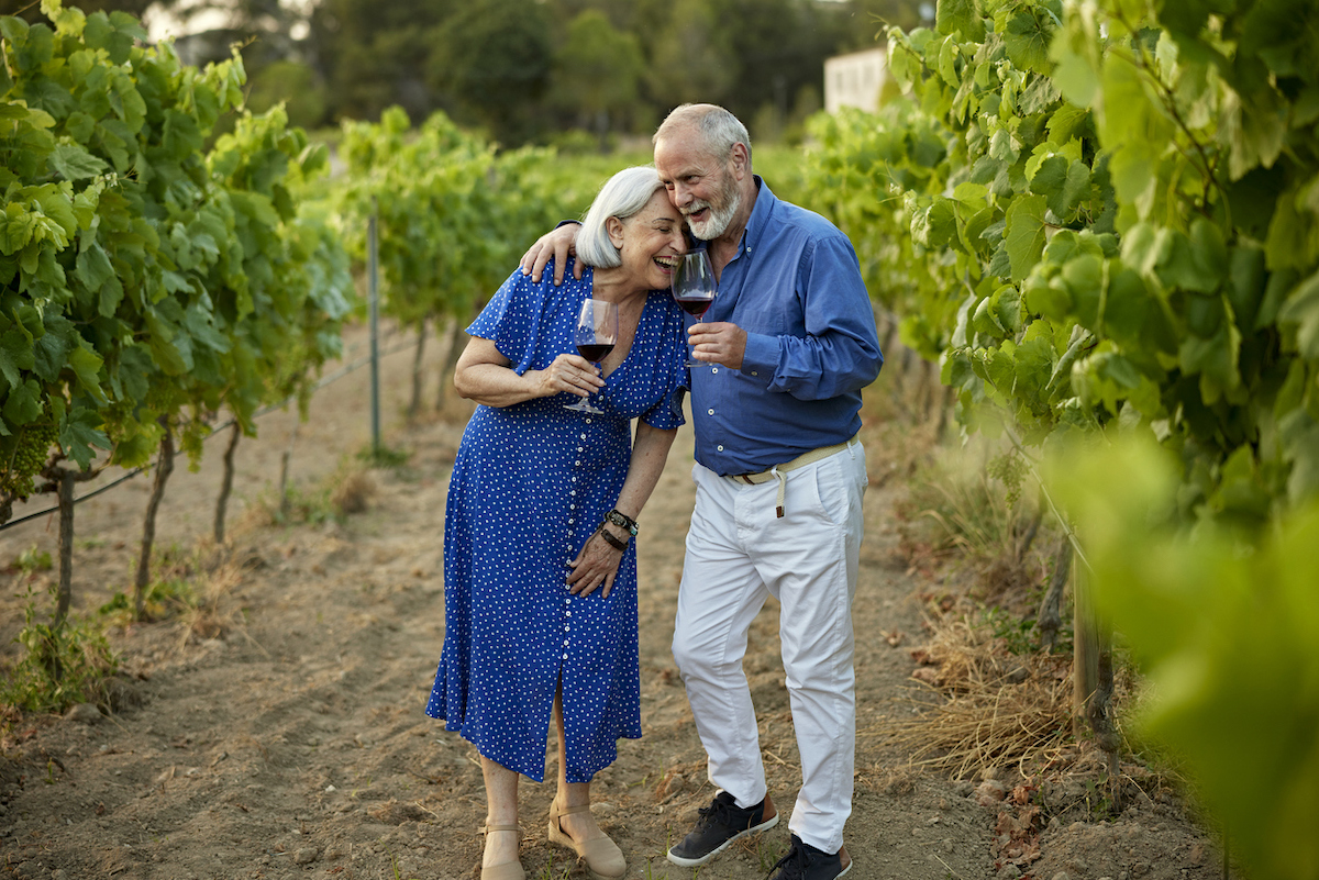 Senior couple, both wearing blue, hugging and laughing while drinking red wine in a vineyard.