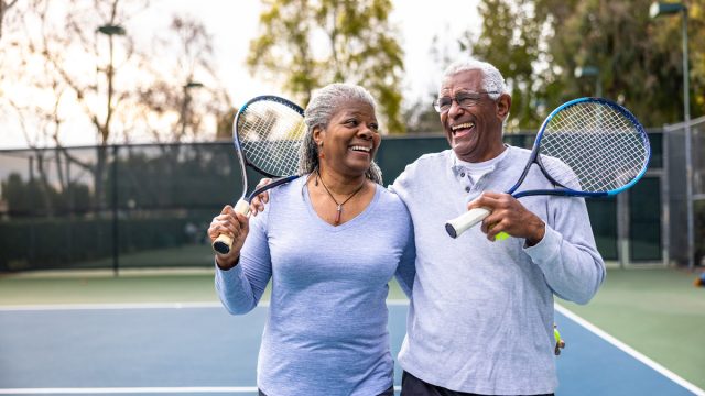 A senior black couple laughing and smiling, leaving the tennis court after their workout.
