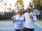 A senior black couple laughing and smiling, leaving the tennis court after their workout.