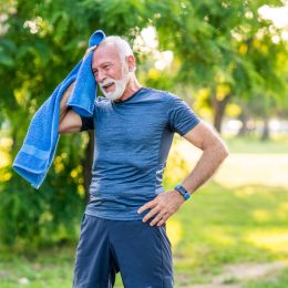 Senior man with white hair and beard wearing blue workout clothes takes a break while exercising outside on a hot day, wiping his face with a blue towel