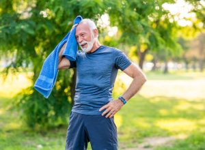 Senior man with white hair and beard wearing blue workout clothes takes a break while exercising outside on a hot day, wiping his face with a blue towel