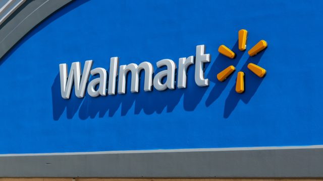 Horizontal, medium closeup of "Walmart" superstore's exterior facade brand and logo signage in white letters against a reflective, blue background and yellow logo