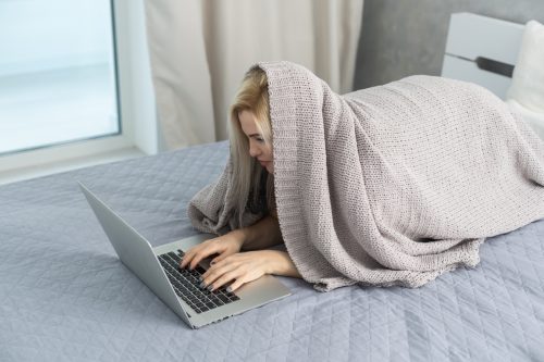 woman writing funny 'out of office' messages from bed while hiding under a blanket