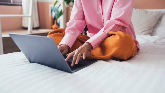 Woman on bed on laptop pink shirt orange pants