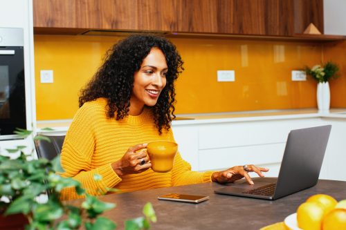Portrait of a woman with black curly hair smiling while using laptop computer in kitchen. She's wearing a yellow-orange sweater that matches the wall color