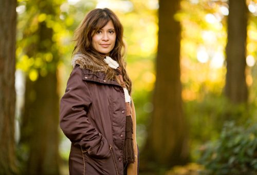 Woman outside surrounded by trees wearing a brown jacket and scarf
