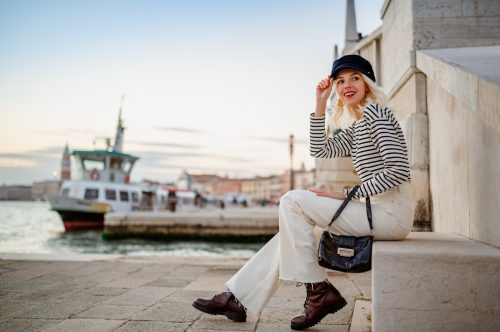 Elegant happy smiling fashionable woman wearing trendy baker boy cap, striped long sleeve shirt, white flared jeans, boots, holding small bag, posing near bridge in Venice.